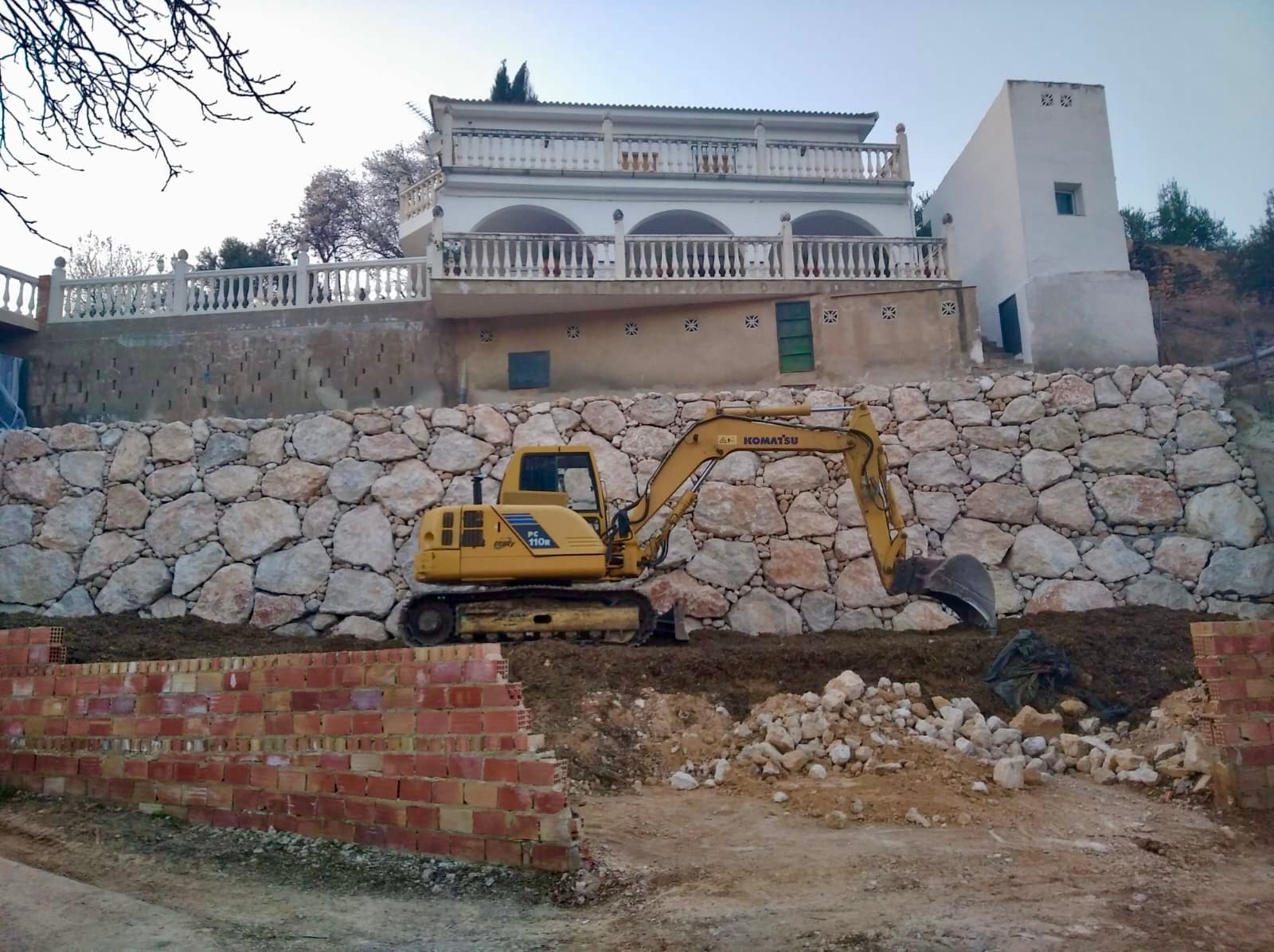 Excavadora amarilla Komatsu trabajando en un muro de piedra frente a una casa blanca.