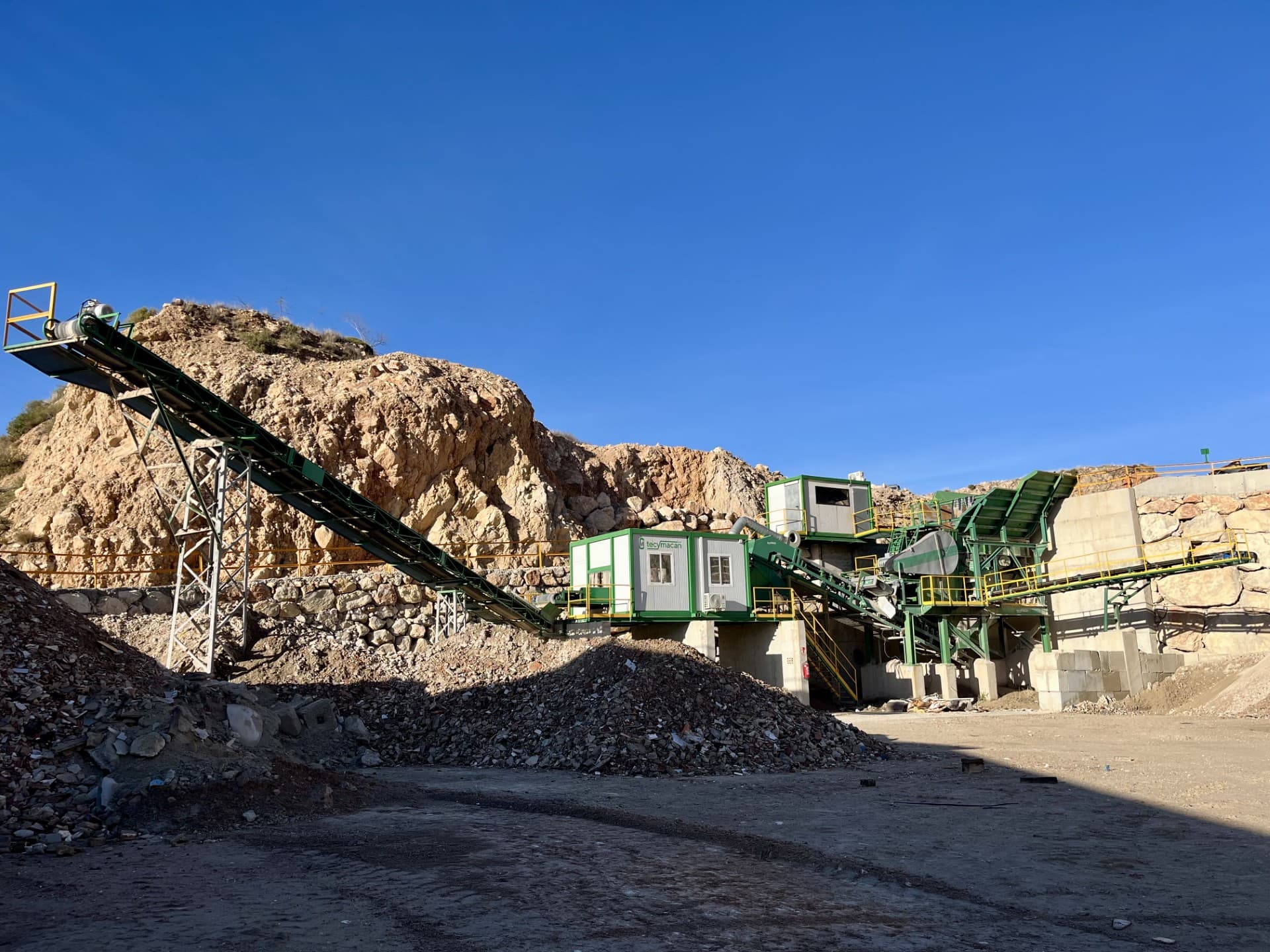 Planta de trituración y cinta transportadora en una cantera bajo un cielo azul despejado.