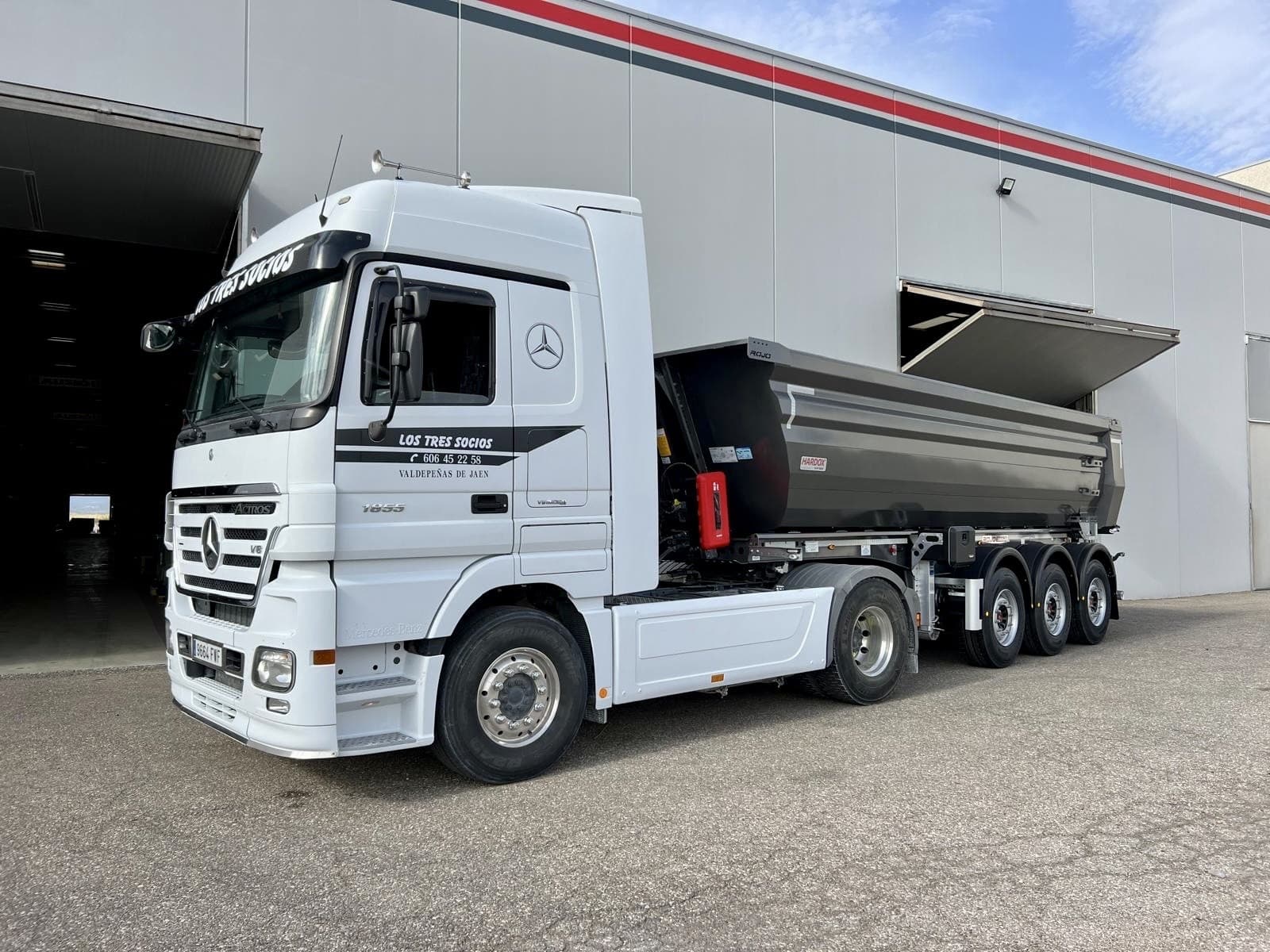 Camión Mercedes-Benz blanco con remolque basculante negro estacionado frente a un edificio industrial moderno.