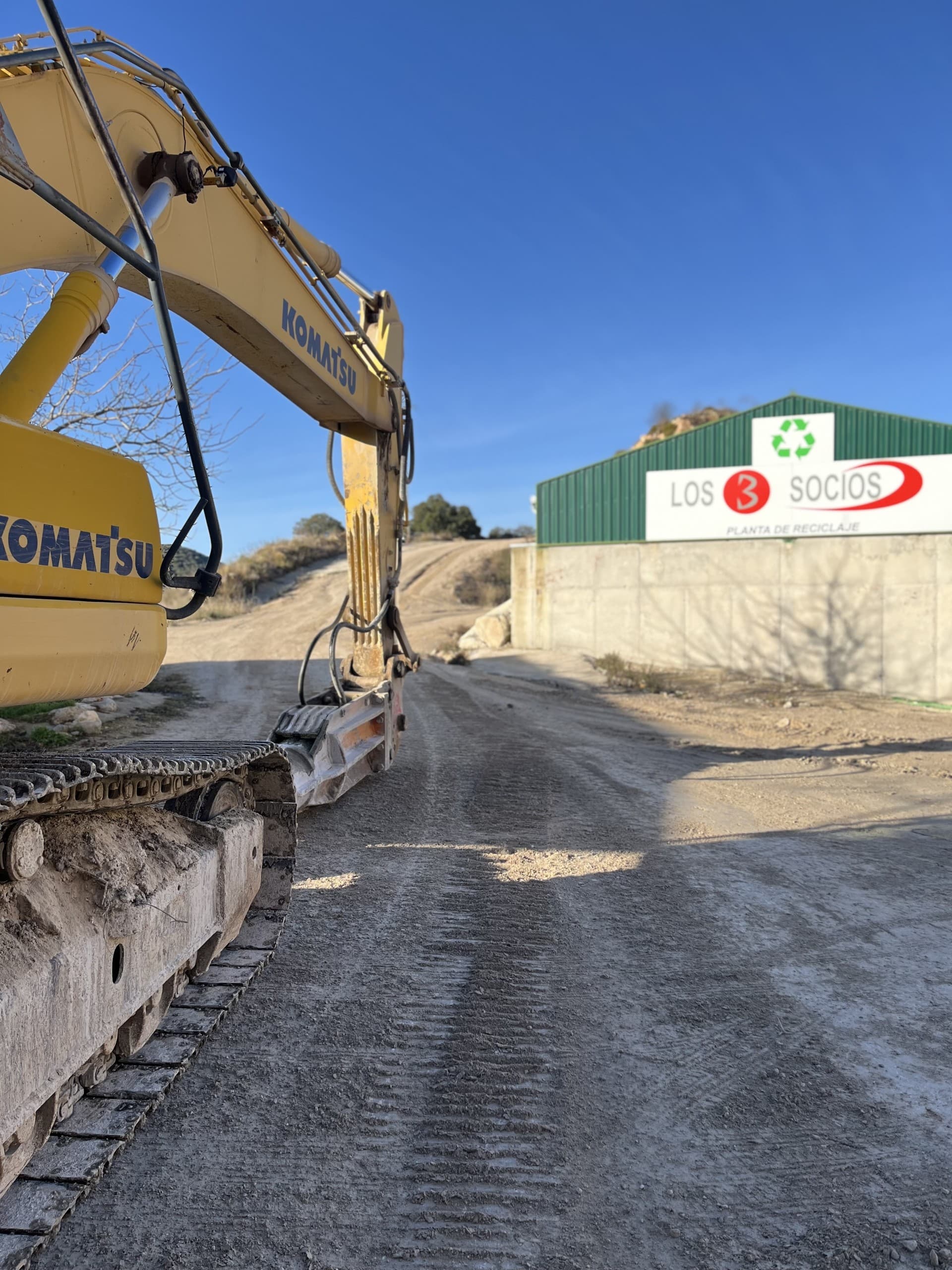 Excavadora Komatsu amarilla en un camino de tierra frente a una planta de reciclaje industrial.