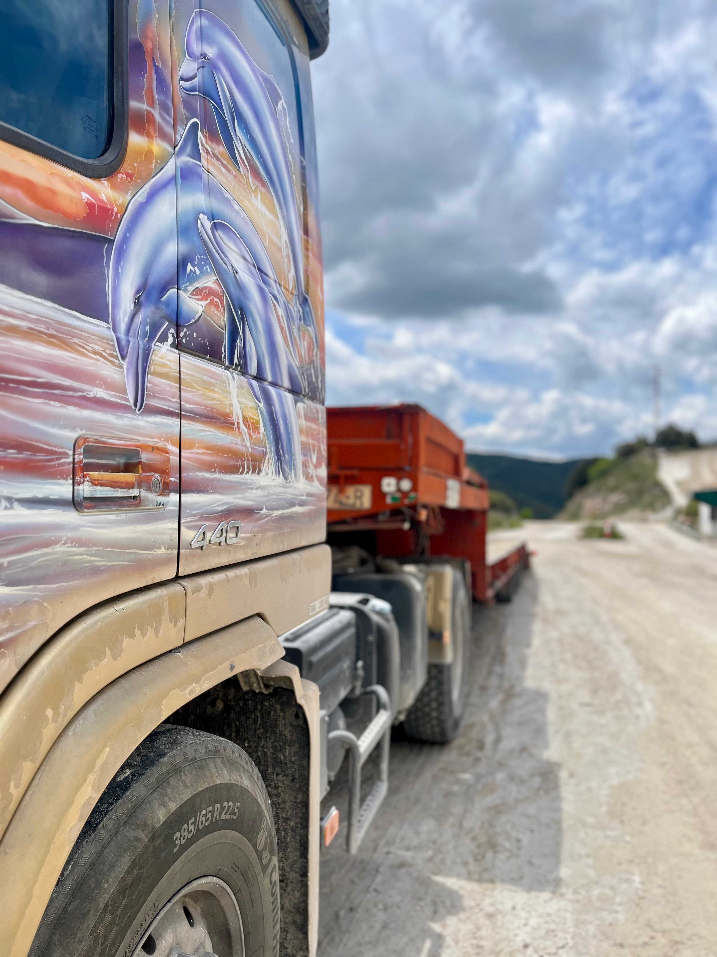 Camión decorado con delfines saltando, remolcando una plataforma roja por un camino de tierra.