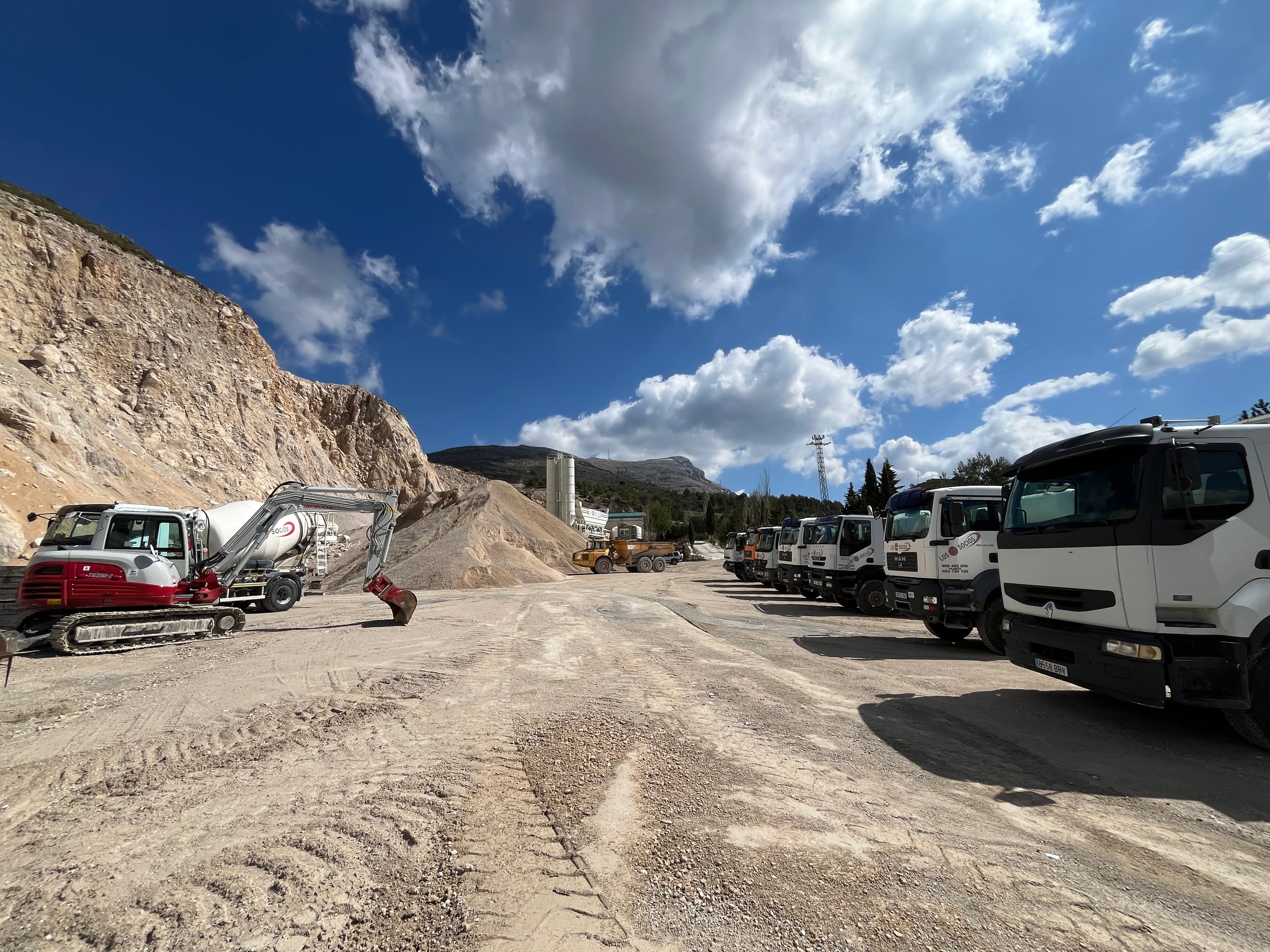 Excavadora y camiones blancos en una cantera de arena bajo un cielo azul nublado.