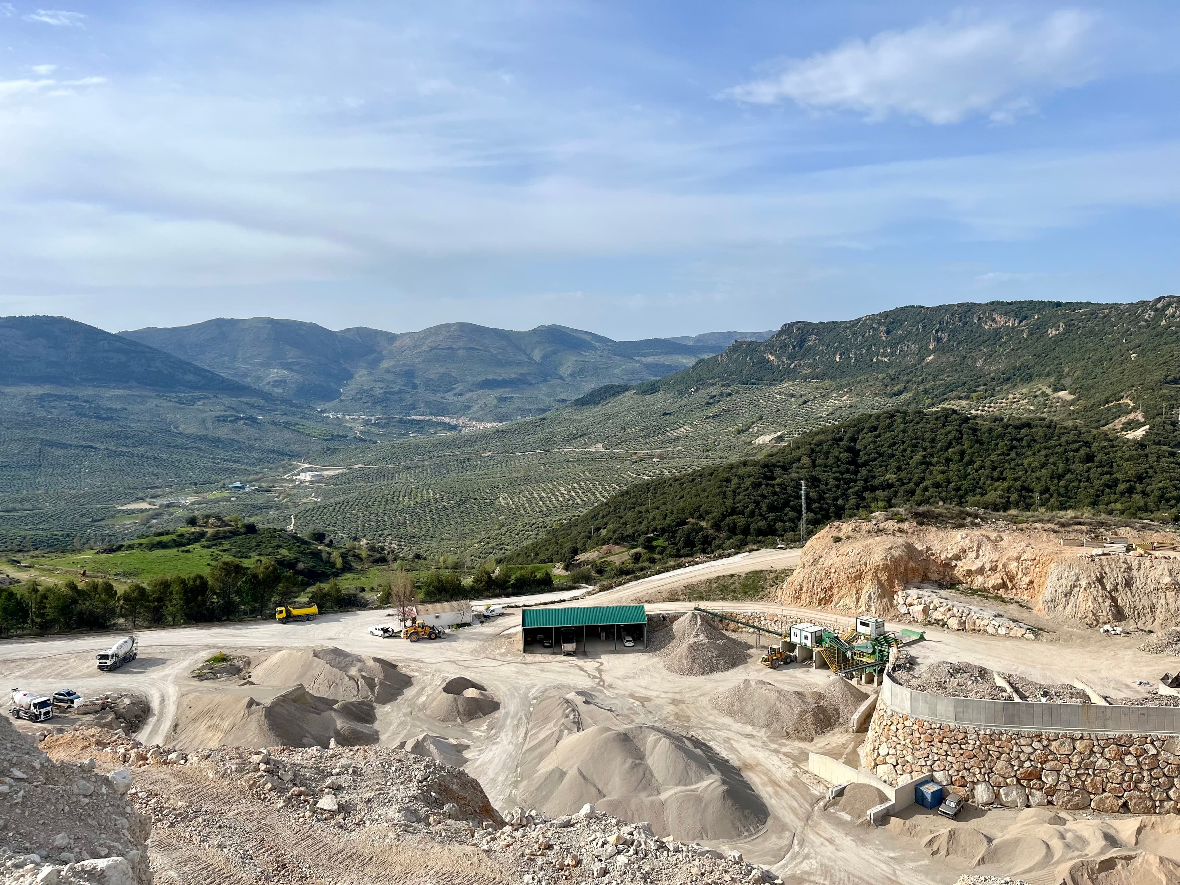 Cantera industrial con maquinaria y montículos de arena frente a un extenso valle de montañas.