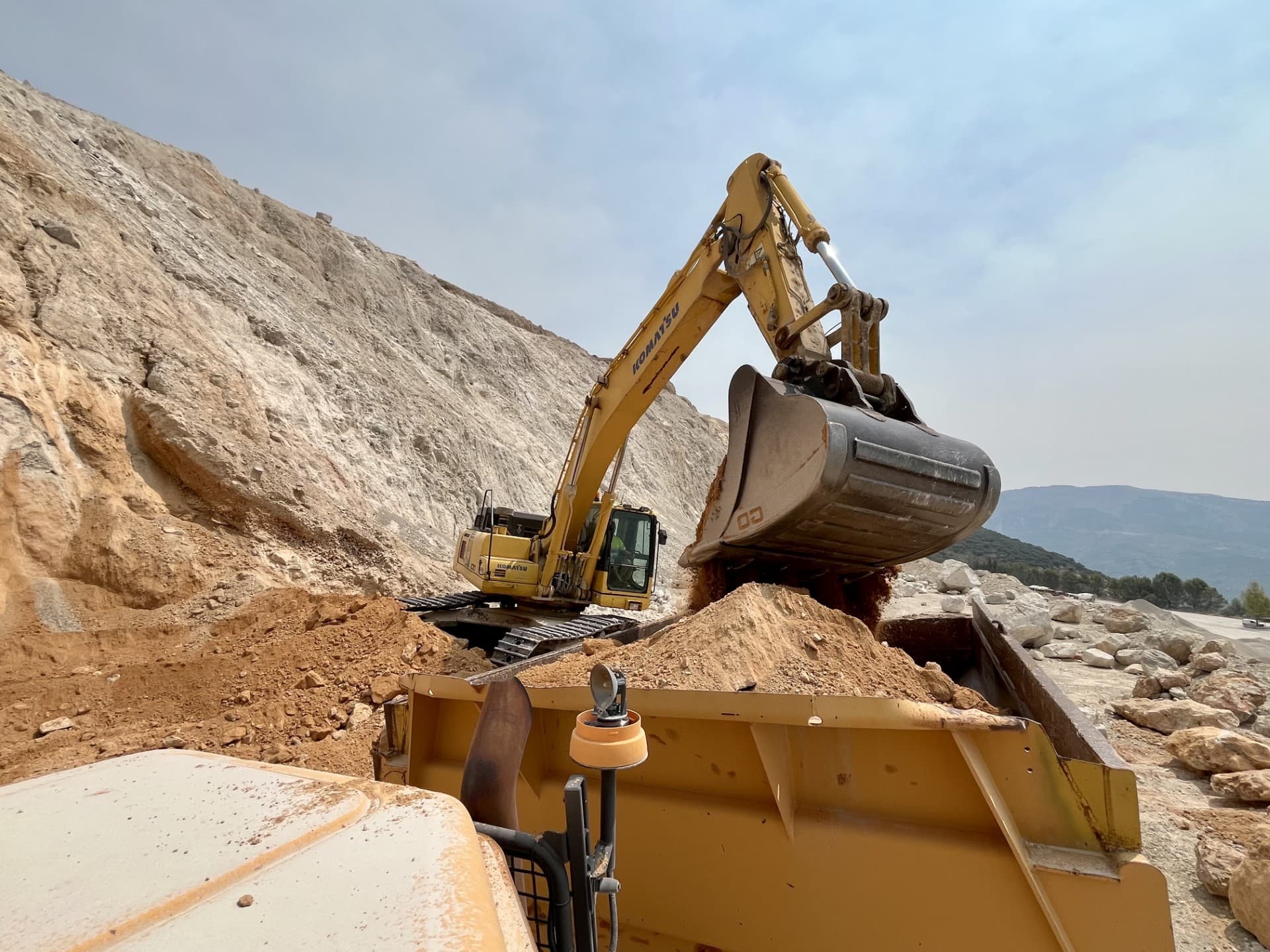 Excavadora amarilla cargando tierra en un camión volquete frente a una gran ladera rocosa.