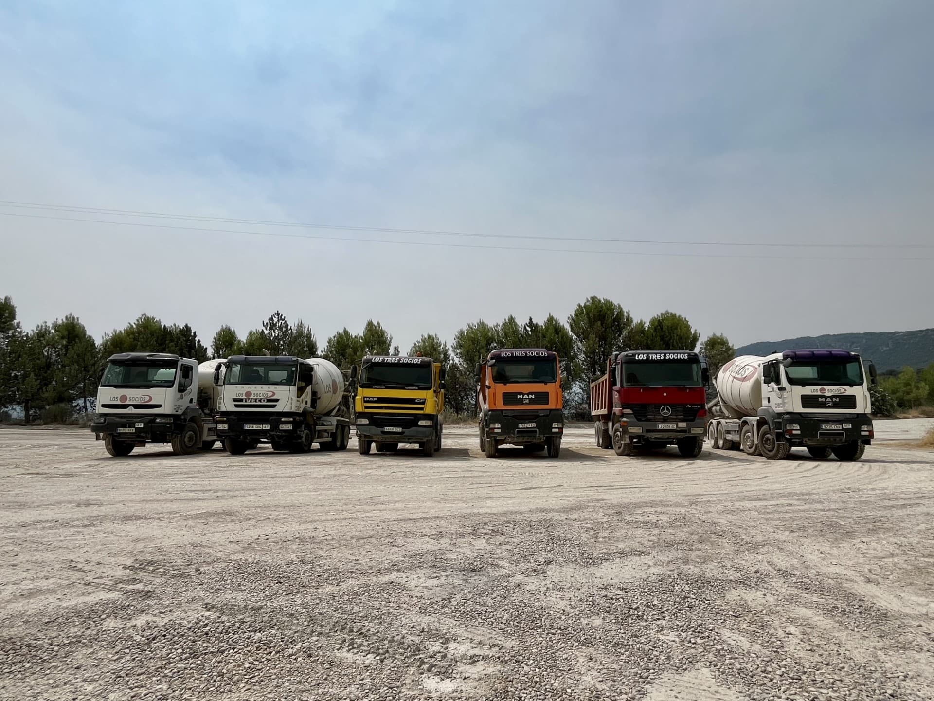 Seis camiones de construcción de Los Tres Socios alineados en un terreno de grava.
