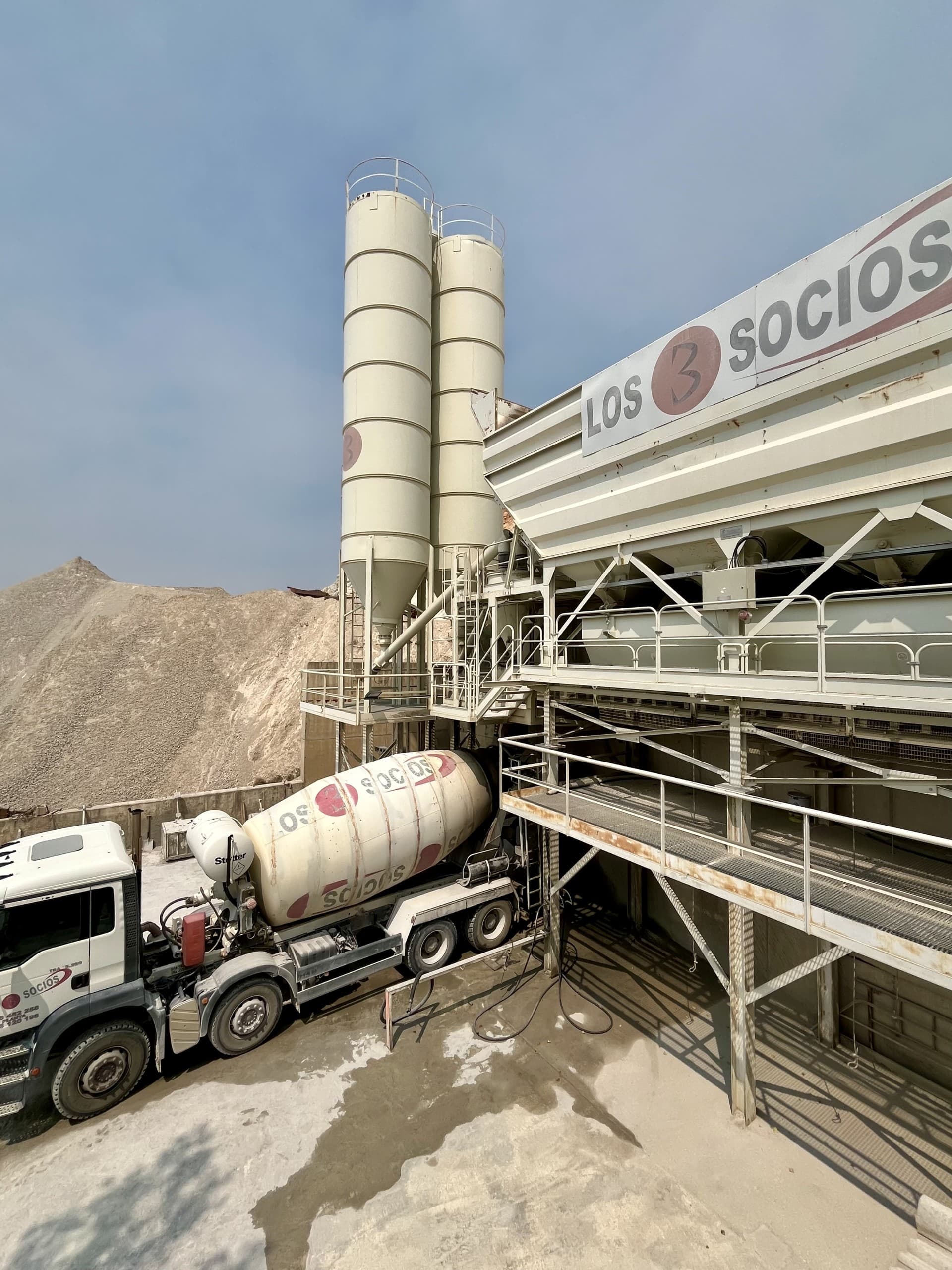 Camión hormigonera de Los 3 Socios estacionado en una planta de concreto con silos.