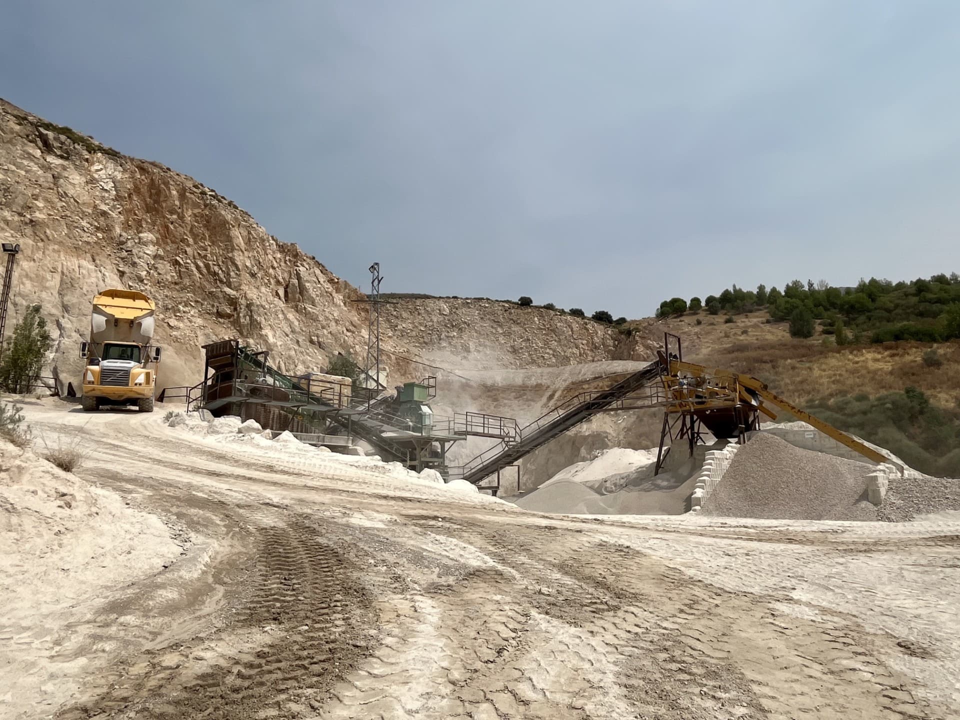 Camión volquete amarillo junto a cintas transportadoras en una cantera de piedra caliza polvorienta.
